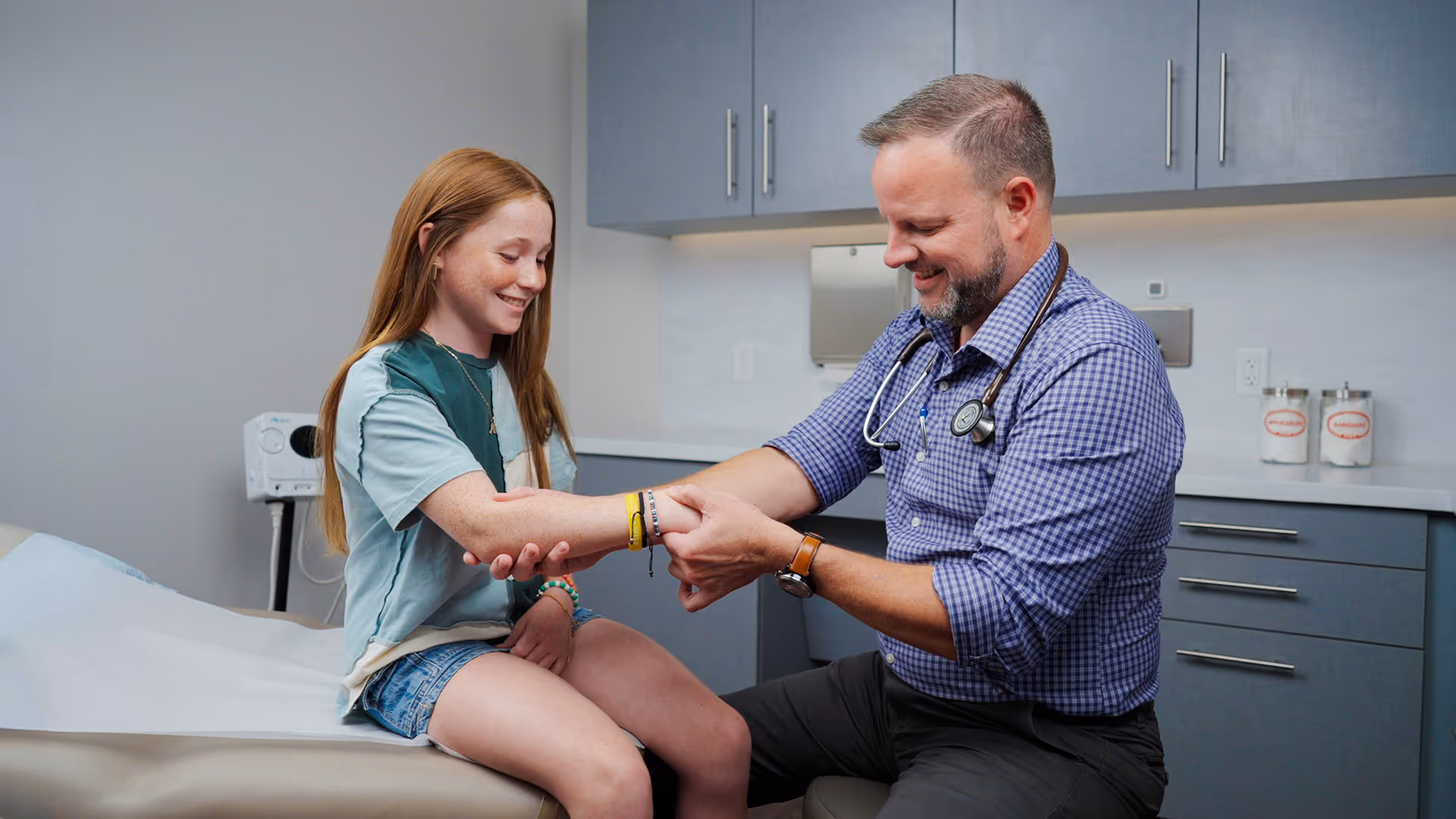 Doctor with stethoscope examining the arm of a smiling young girl sitting on an exam table in a medical office.