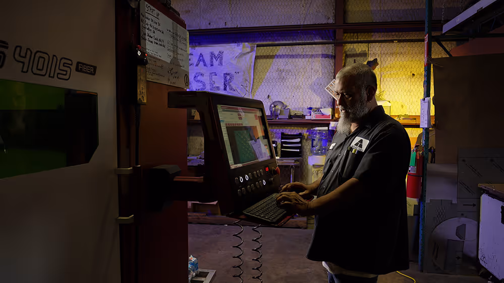 Man with grey beard at CNC machine, blue and yellow colored lights in the background