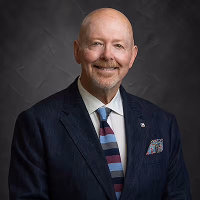 Smiling bald man with a goatee wearing a dark suit, white shirt, striped tie, and a patterned pocket square against a dark textured background.