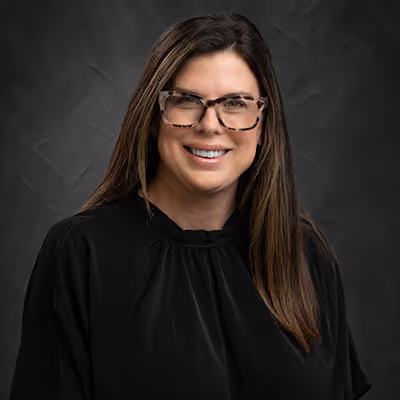 Smiling woman with long brown hair wearing tortoiseshell glasses and a black top against a dark textured background.