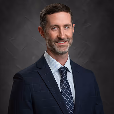 Professional man with short dark hair and beard wearing a navy blue suit, checkered shirt, and patterned tie against a dark textured background.