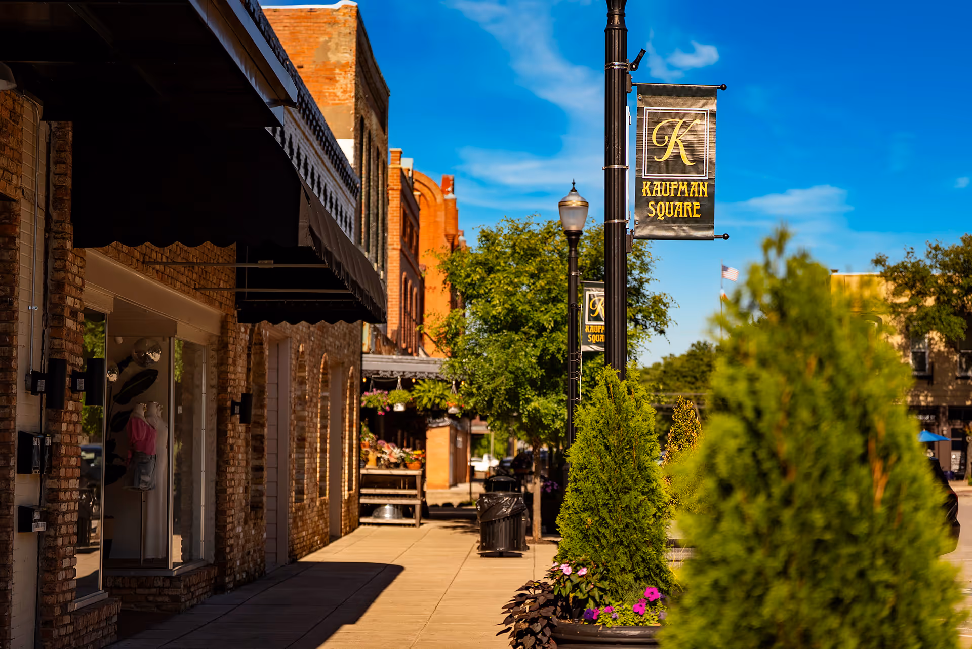 Sunny street view of brick buildings and shops with black awnings, green trees, and Kaufman Square banners on lampposts.