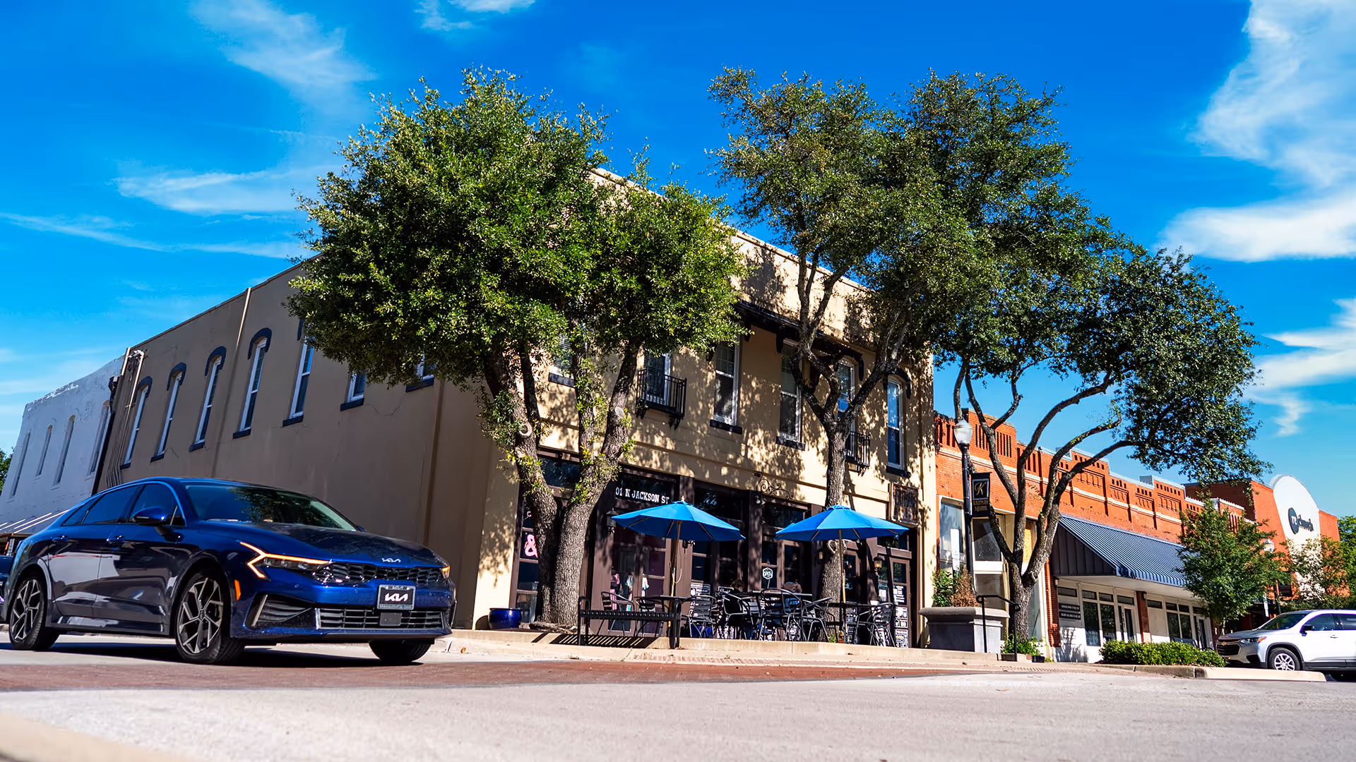 Street view of a small town with trees, outdoor seating under blue umbrellas, and parked cars under a bright blue sky.