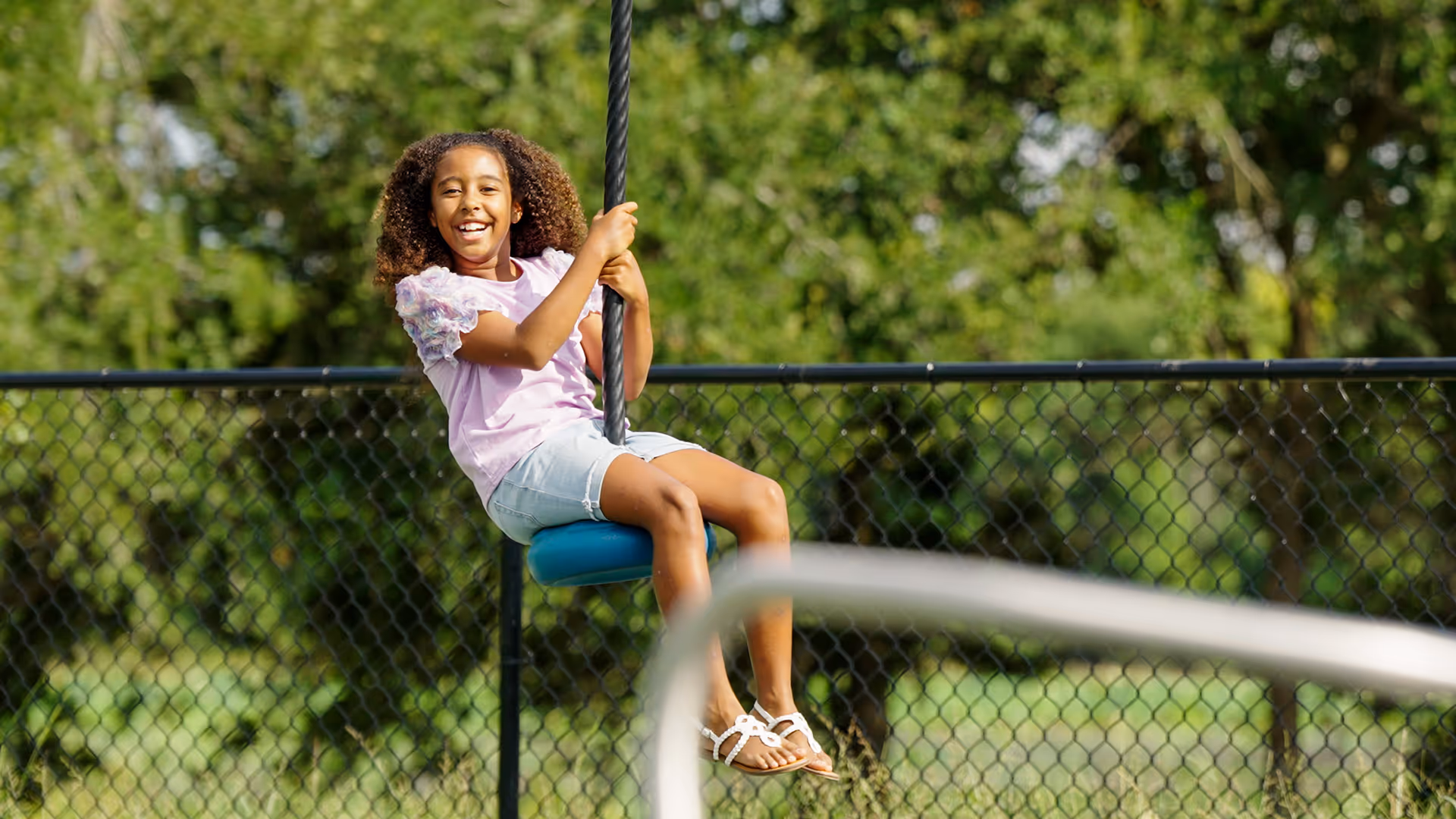 Smiling girl wearing a lavender shirt and shorts swinging on a blue rope swing in a park with green trees and a chain-link fence in the background.