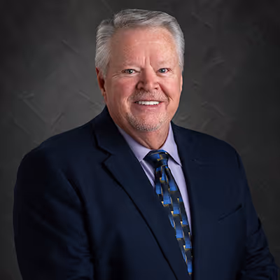 Smiling older man with gray hair wearing a dark blazer, patterned tie, and light purple shirt.