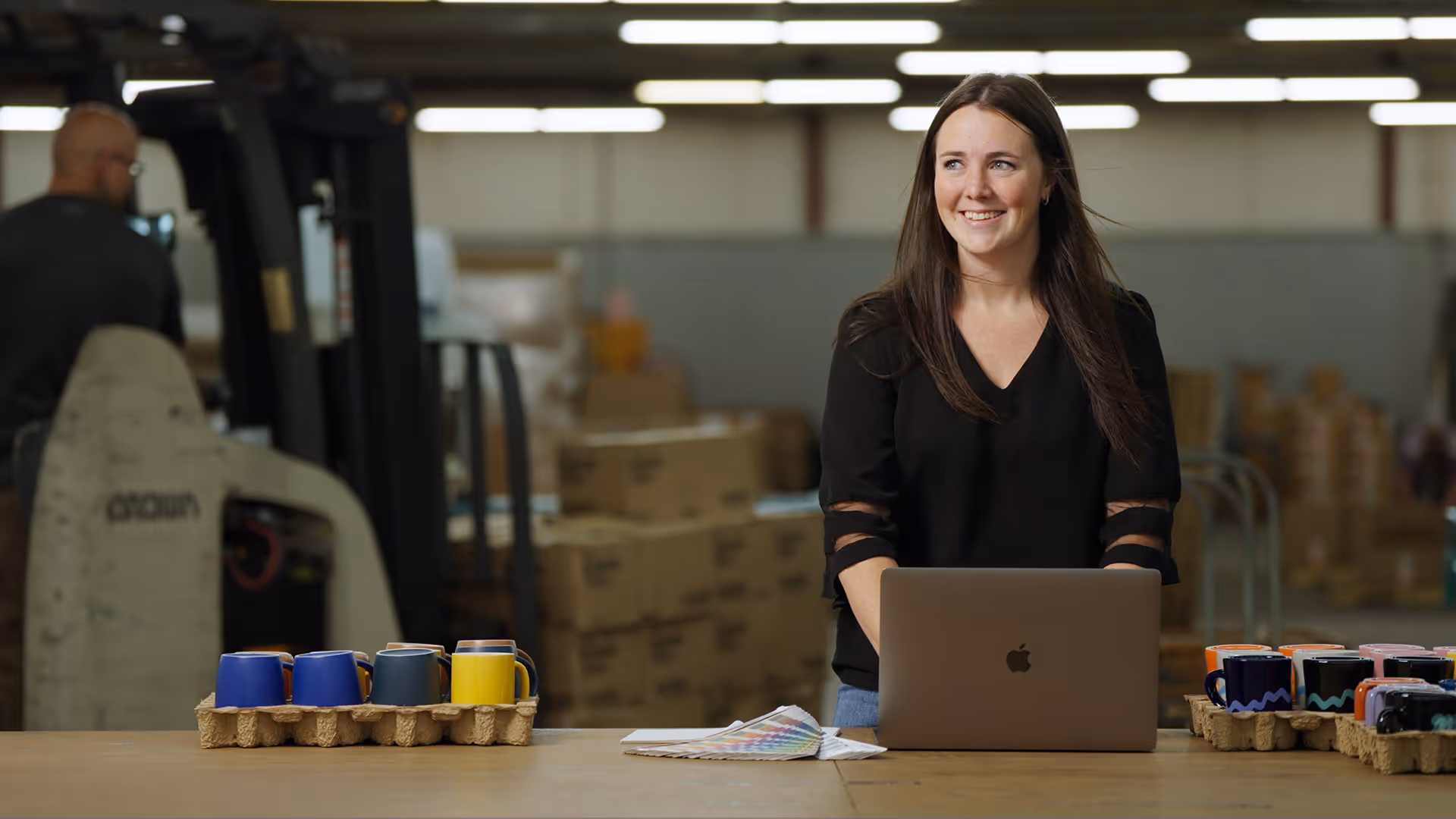 Smiling woman with long brown hair using a laptop at a table in a warehouse with trays of colorful mugs and a color swatch book.