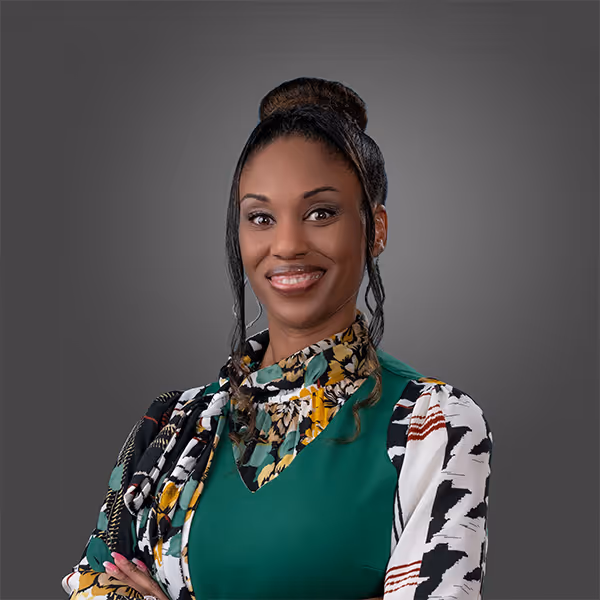 Smiling woman with styled hair in a bun wearing a green dress with a floral and patterned scarf on a gray background.