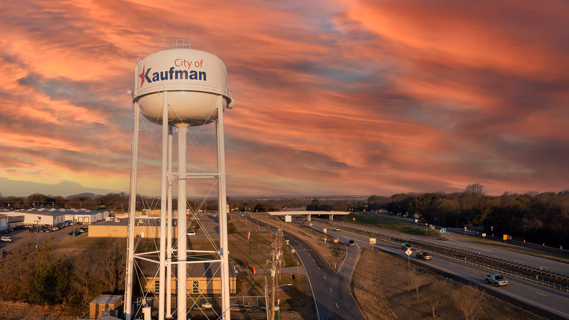 White City of Kaufman water tower near highway at sunset with orange and purple clouds.