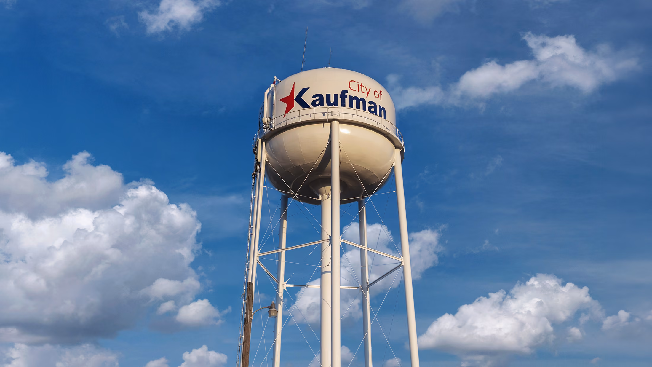 Tall white water tower with City of Kaufman logo against a partly cloudy blue sky.
