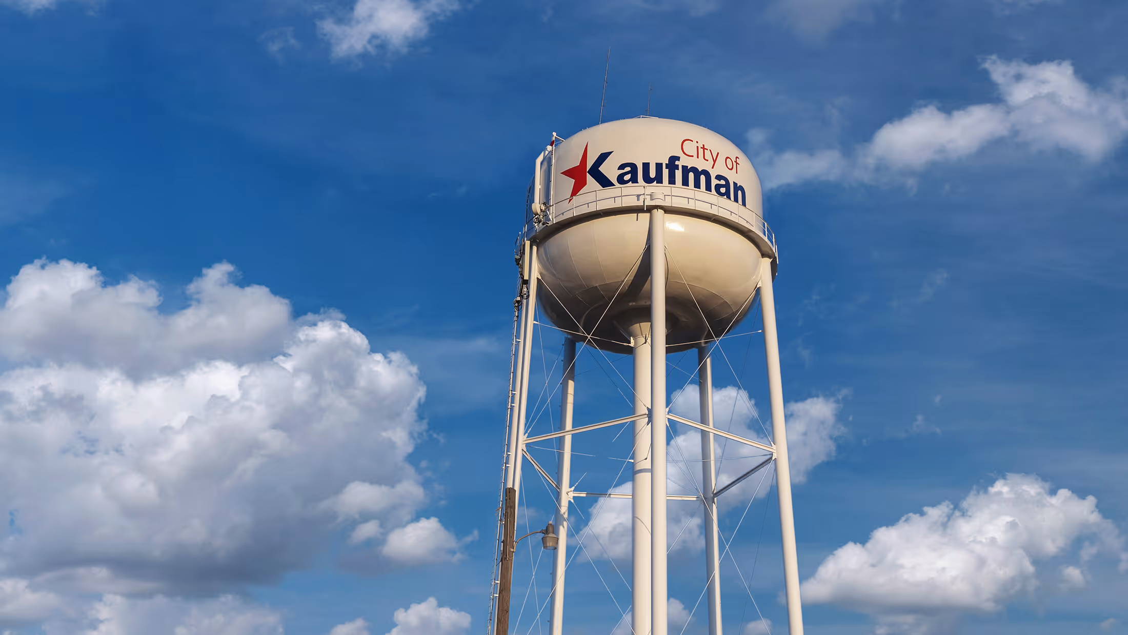 Tall white water tower with City of Kaufman logo against a partly cloudy blue sky.