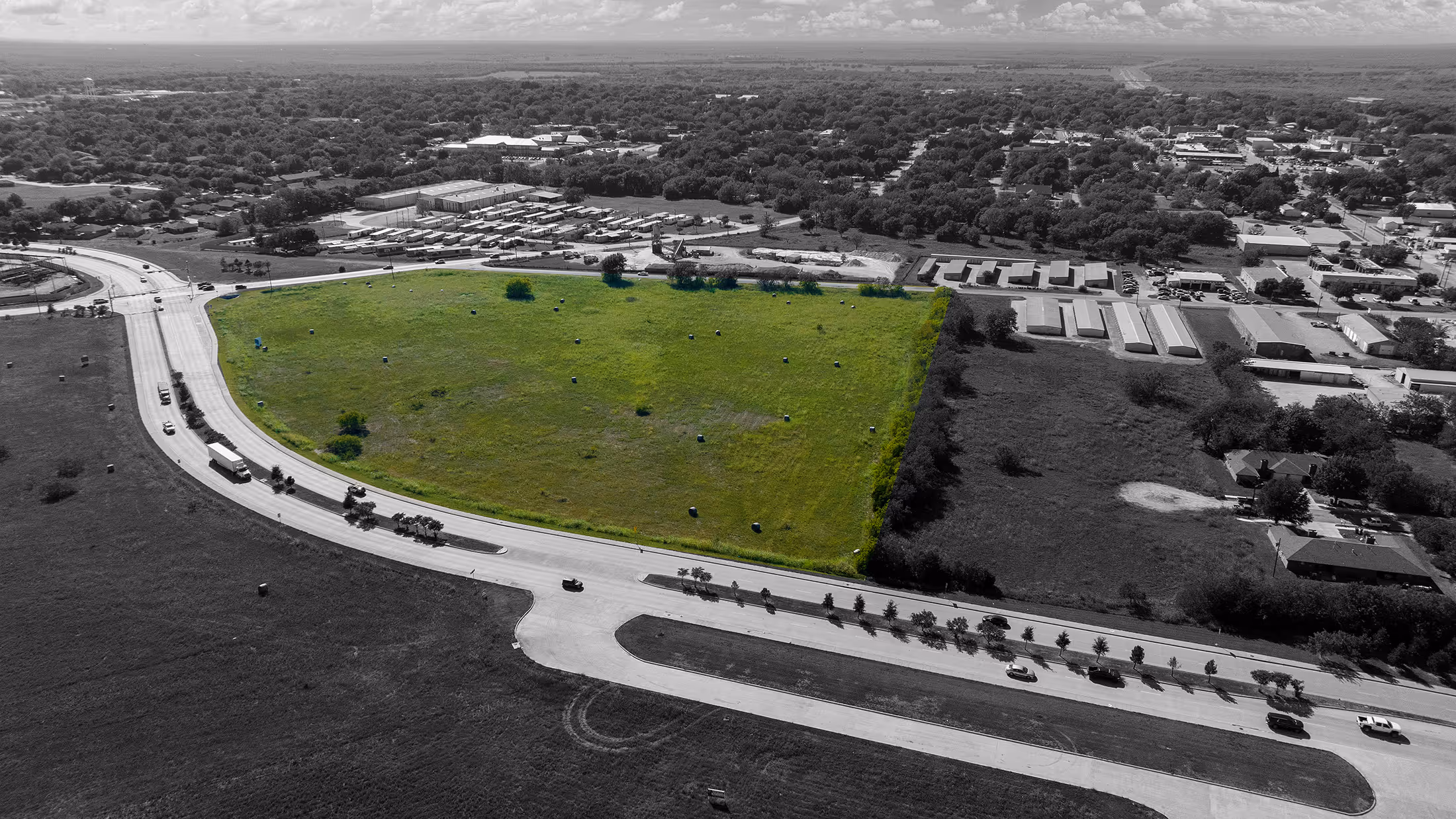 Aerial view of a green field bordered by roads with buildings and trees in the background.