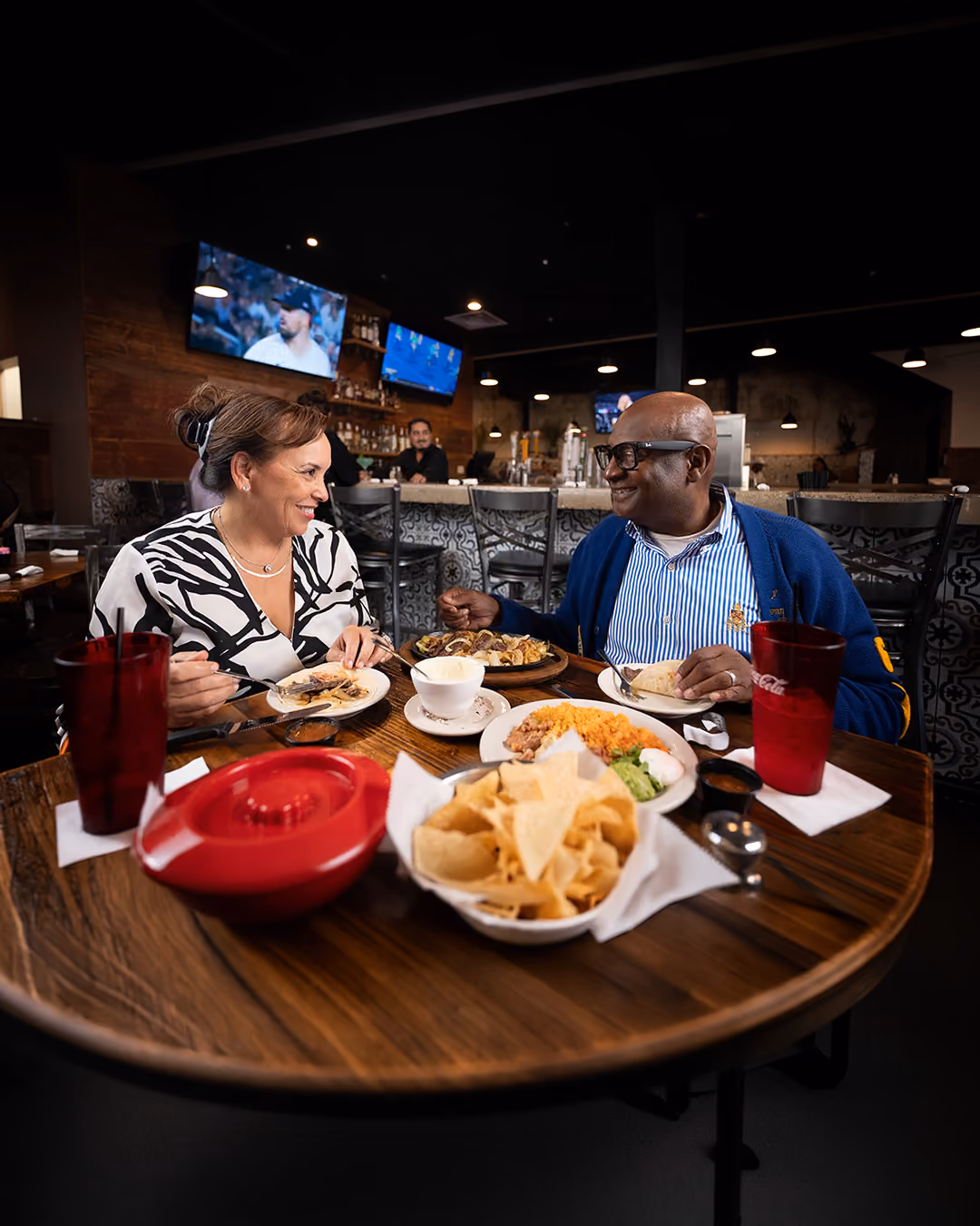 Smiling man and woman eating Mexican food with chips, salsa, and drinks at a restaurant table.