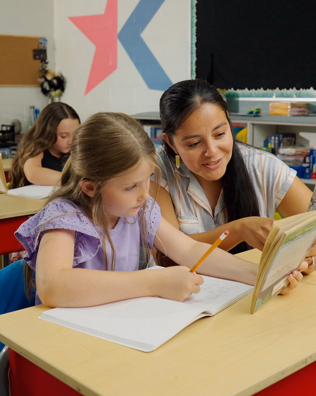 Teacher helping young girl with reading and writing at classroom desk.
