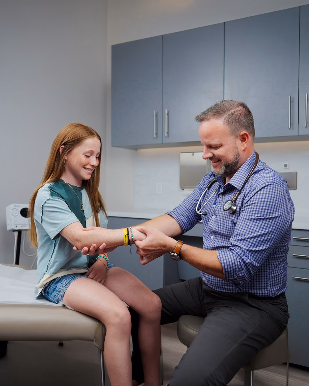 Smiling male doctor examines the arm of a young girl sitting on an exam table in a medical office.