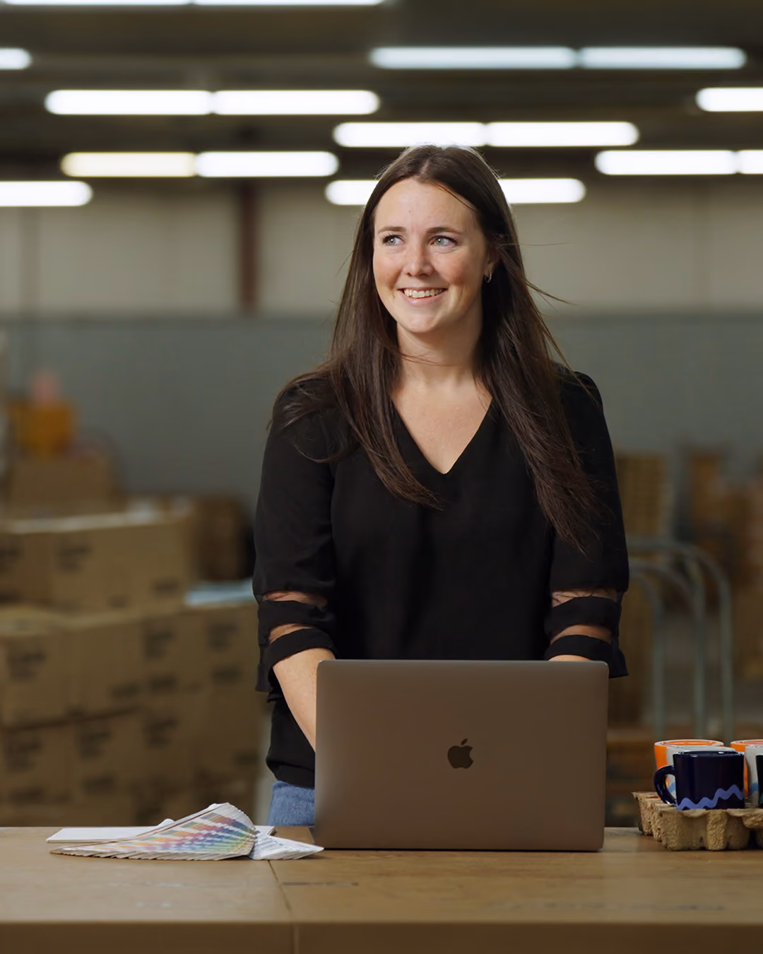Smiling woman in black top standing behind a laptop with a color swatch and mugs on a table.
