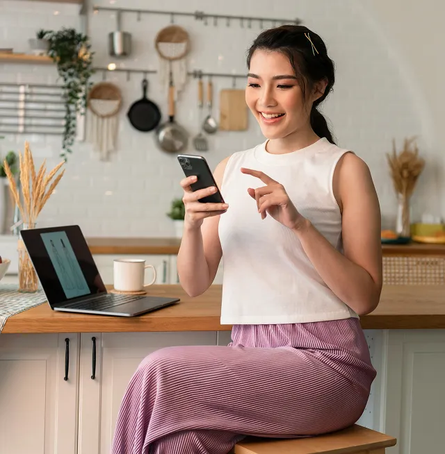 Smiling woman sitting on a stool using a smartphone at a kitchen counter with a laptop and coffee mug.