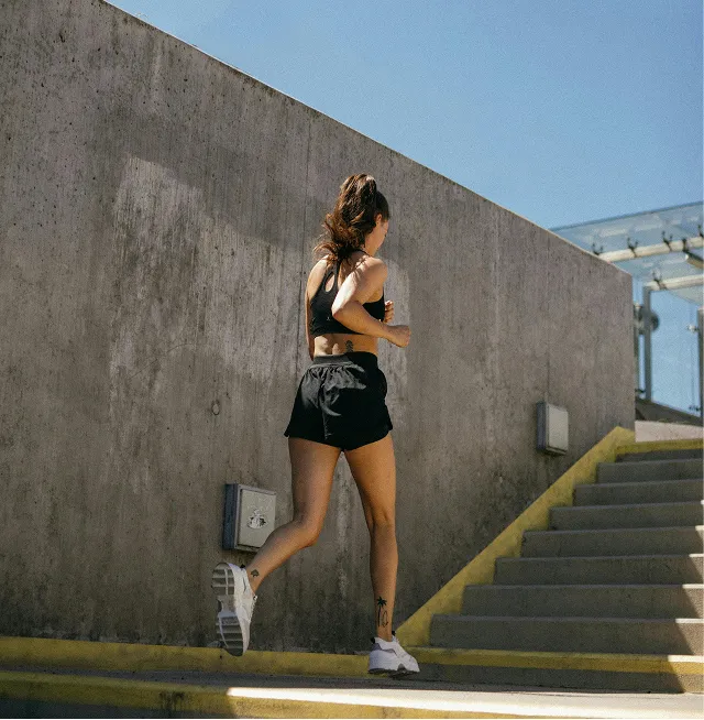 Woman in black sportswear running outdoors past concrete wall and stairs on a sunny day.