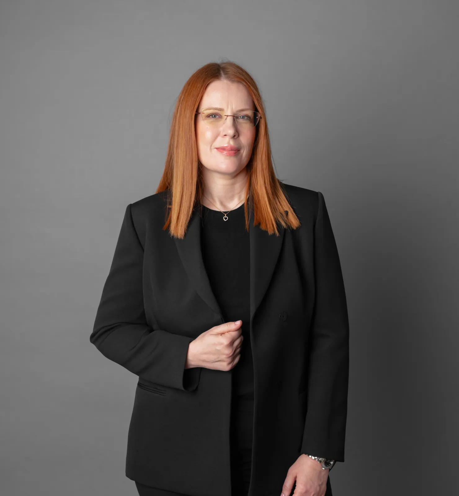 Woman with straight red hair wearing glasses, a black blazer, and a heart-shaped necklace, standing against a gray background.