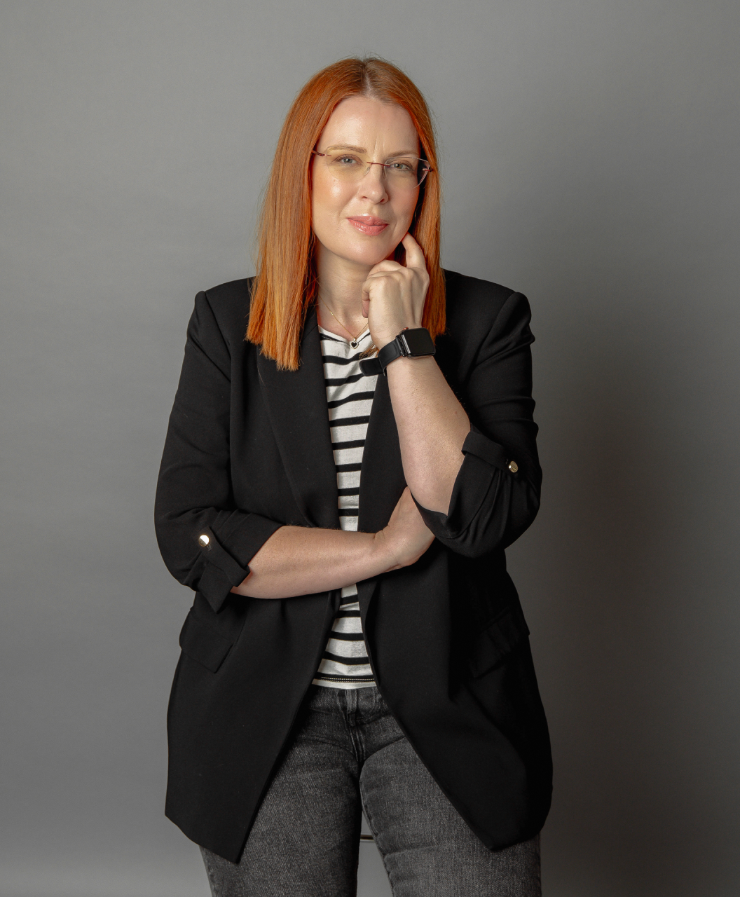 Woman with straight red hair wearing glasses, a black blazer, striped shirt, and smartwatch, posing thoughtfully against gray background.