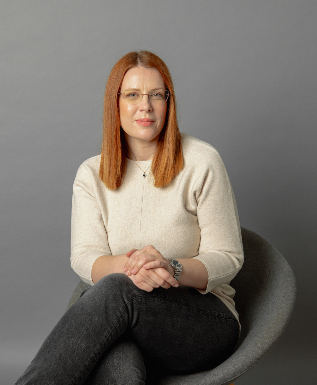 Woman with straight red hair and glasses wearing a light beige sweater and black jeans sitting in a gray chair against a gray background.