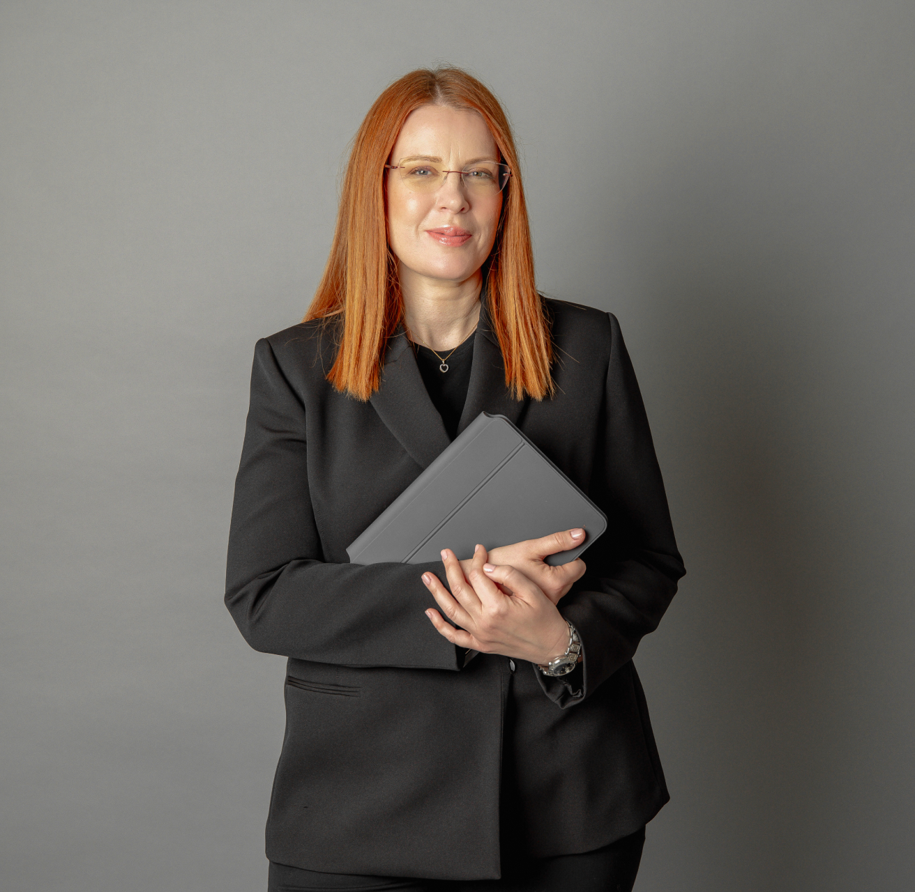 Woman with red hair and glasses wearing a black blazer holding a closed tablet against a gray background.