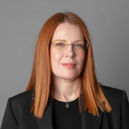 Professional headshot of a woman with straight red hair, glasses, and a black blazer against a gray background.