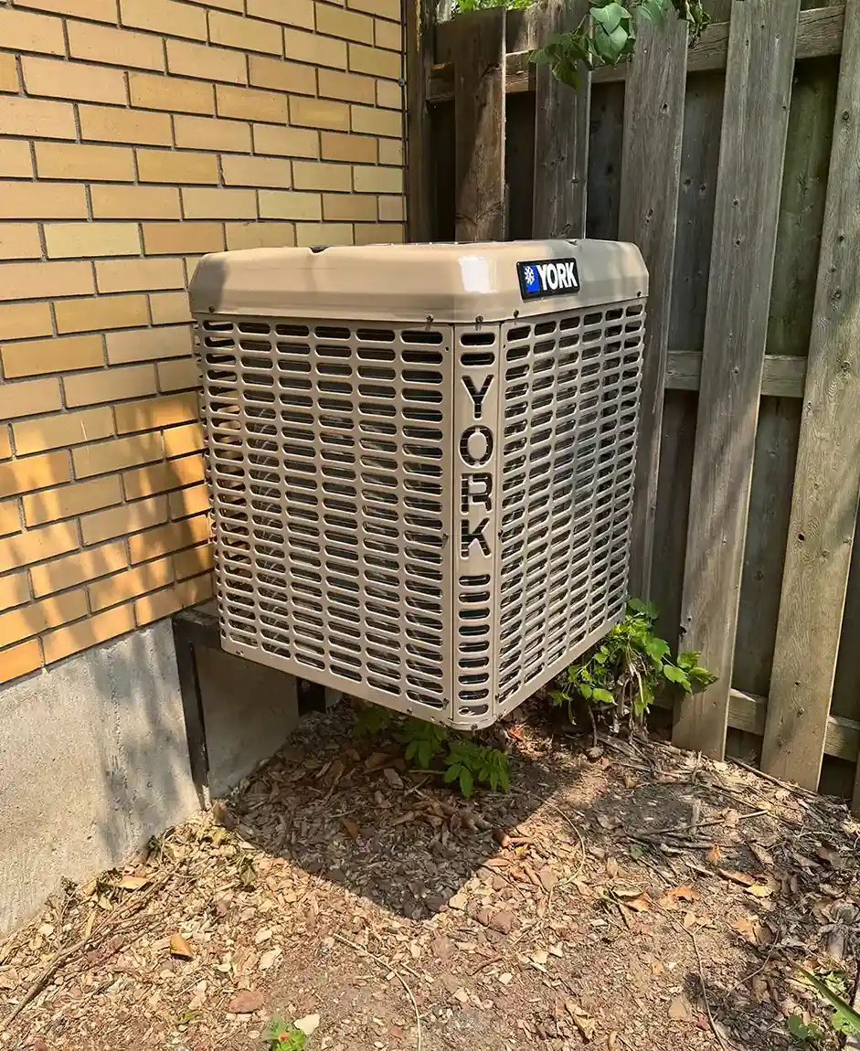 Outdoor YORK air conditioning unit mounted on the side of a house with a brick wall and wooden fence background.