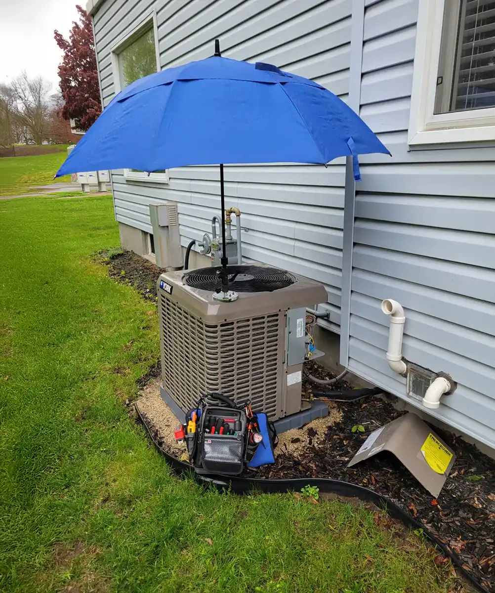 York brand air conditioning unit installed outside a house with a blue umbrella mounted above it and a tool bag placed in front on grass.