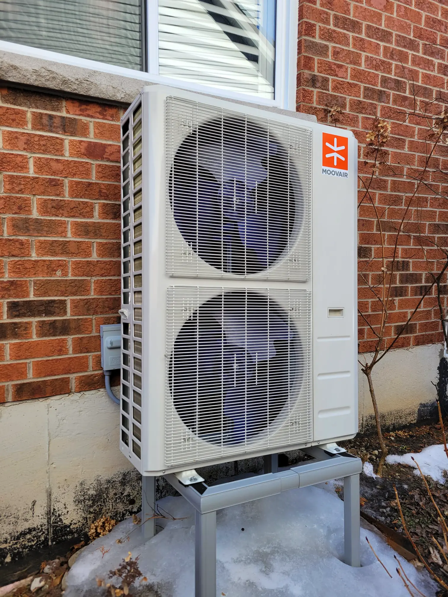 White Moovair heat pump with two large fans mounted on a metal stand outside a brick house on a snowy ground.