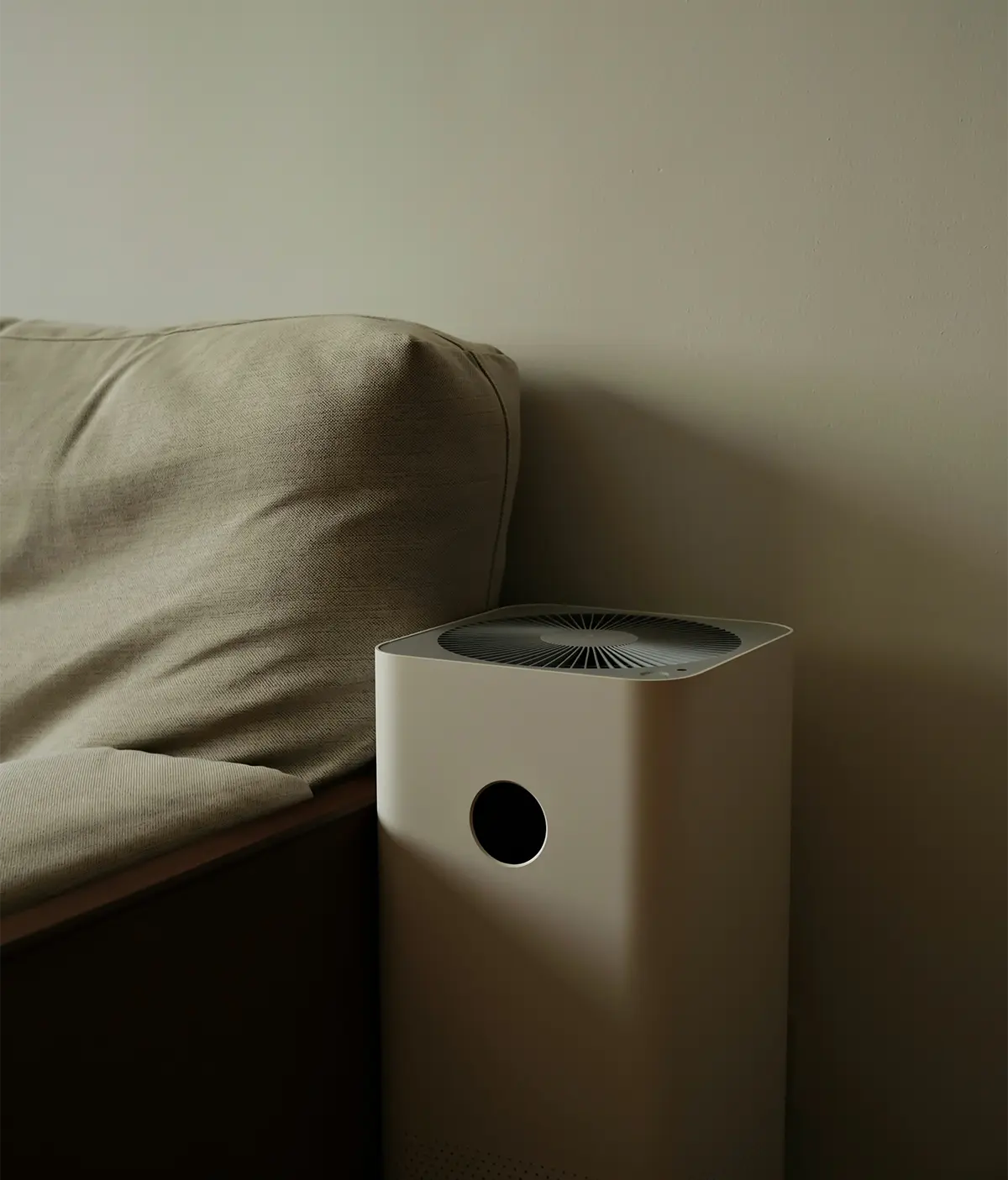 Modern white air purifier placed next to a beige fabric sofa against a plain wall.