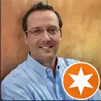 Smiling man with glasses wearing a light blue collared shirt, standing against a blurred warm-toned background.