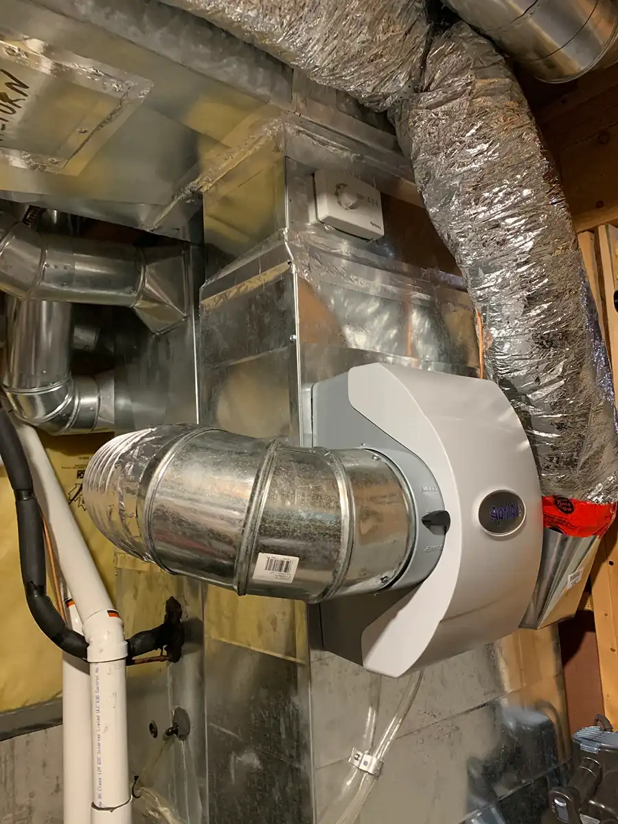 Close-up of HVAC ductwork and an Aprilaire ventilation system installed in a basement with insulated pipes and metallic ducts.