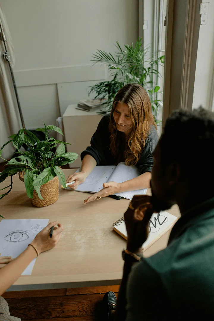 A woman smiling and pointing at a notebook on a table while two people listen and sketch drawings nearby.