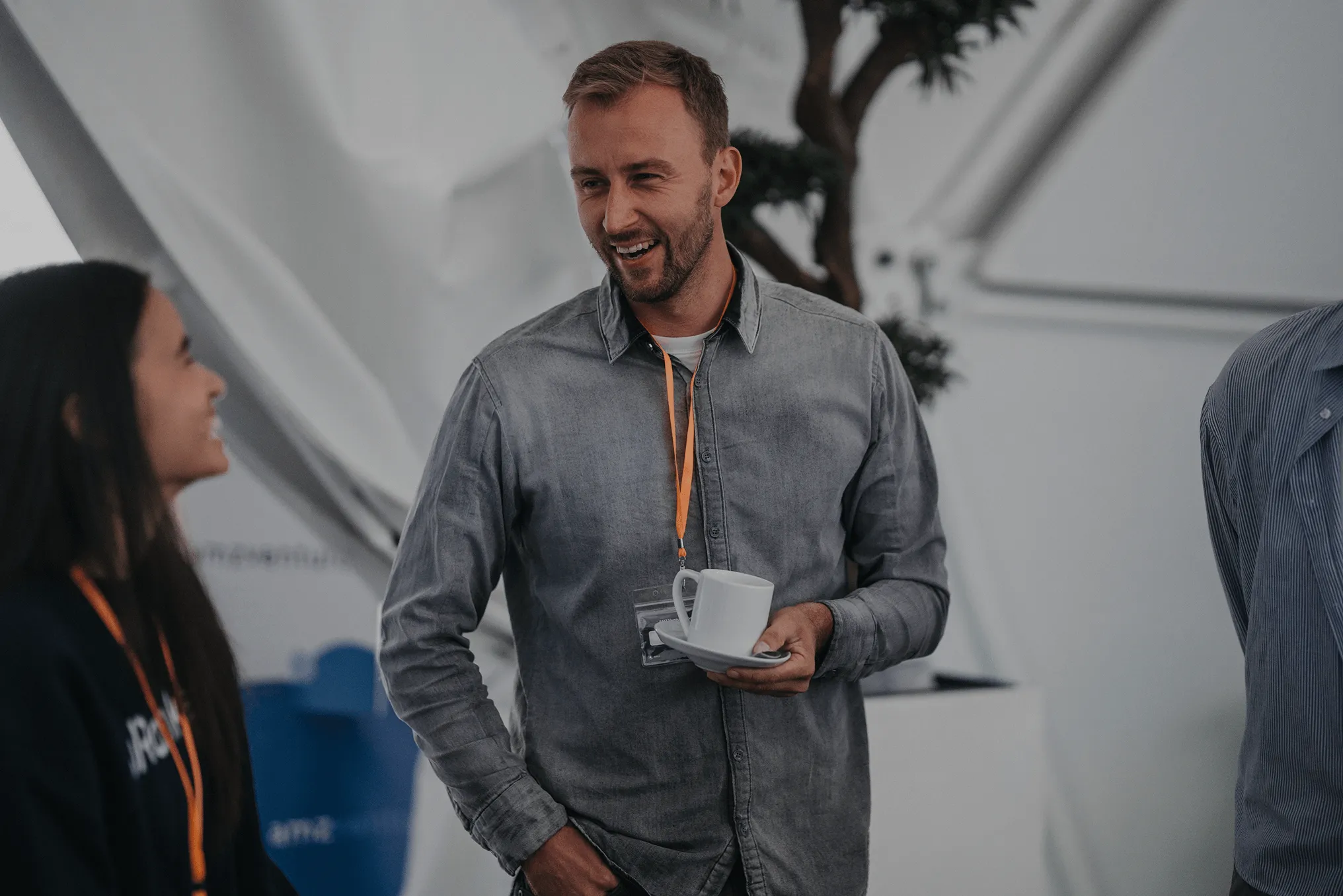 Man in gray shirt holding a white cup and saucer, smiling and talking with a woman wearing an orange lanyard.