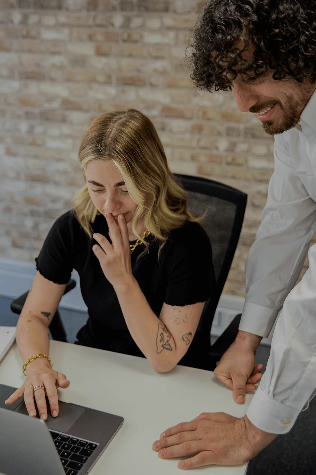 A woman with tattoos and blonde hair is working on a laptop while a man in a white shirt leans over the table beside her, both looking at the screen.