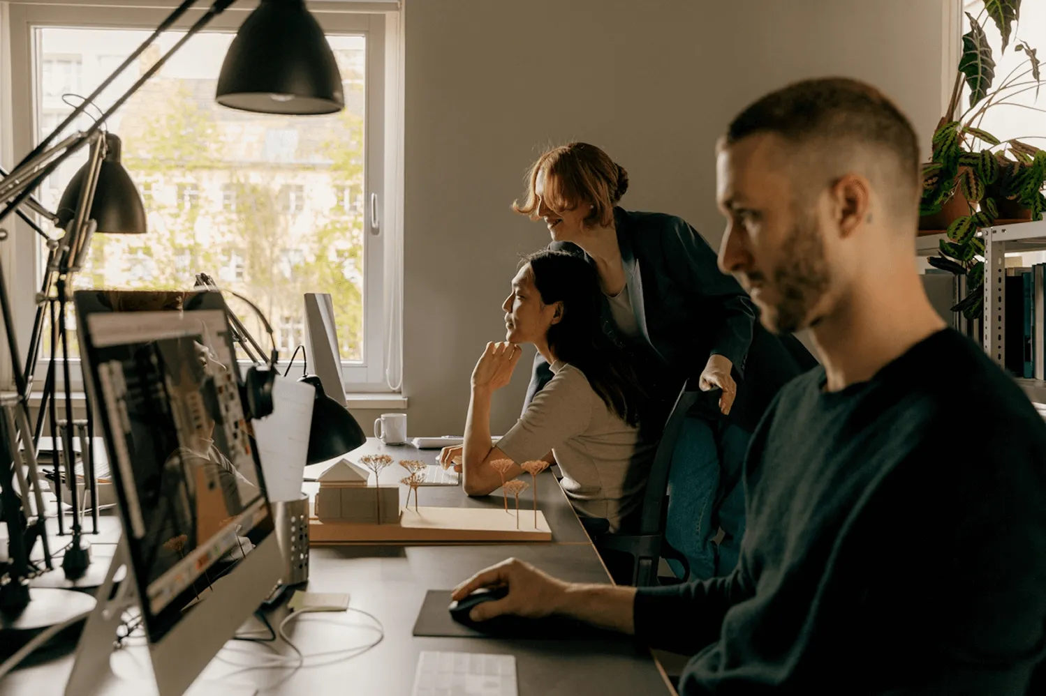 Three coworkers collaborating in a modern office with computers and architectural models on the desk.