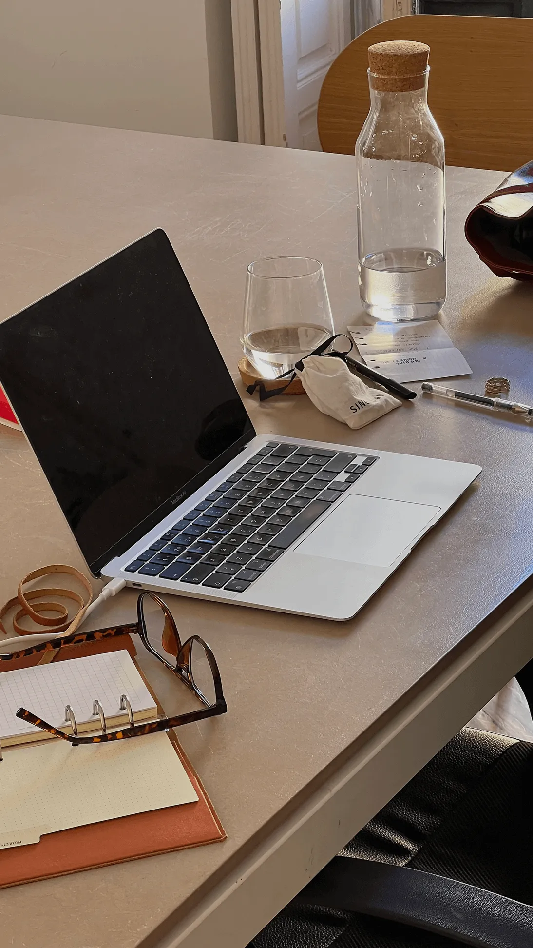 Open laptop on a table next to a glass and bottle of water, reading glasses, notebooks, and writing tools.