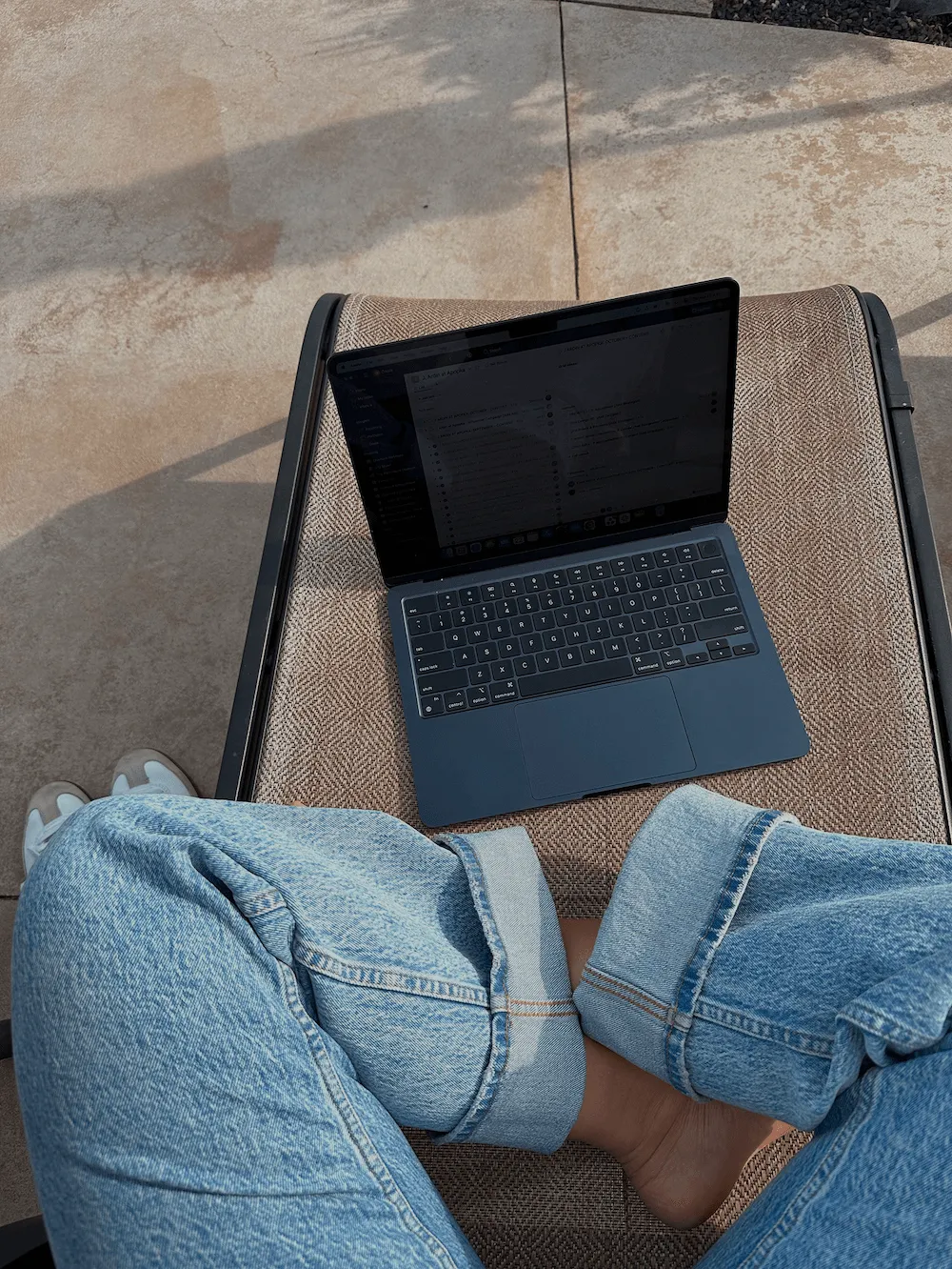 Person sitting outdoors on a lounge chair with crossed legs in rolled-up light blue jeans, a dark laptop resting on the chair beside them.