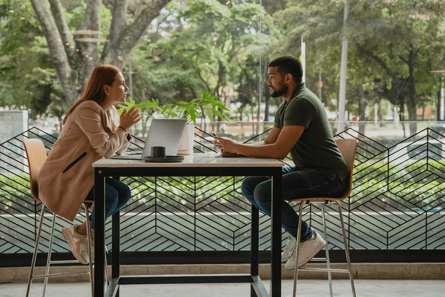 Two people sit across from each other at a table with a laptop and plant between them, engaged in conversation in a modern indoor space with large windows and greenery outside.
