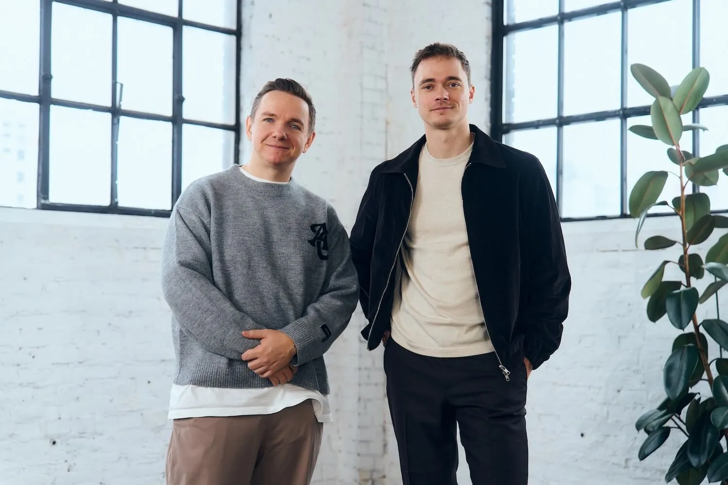 Two men standing indoors against a white brick wall with large windows, one wearing a grey sweater and the other a black jacket.