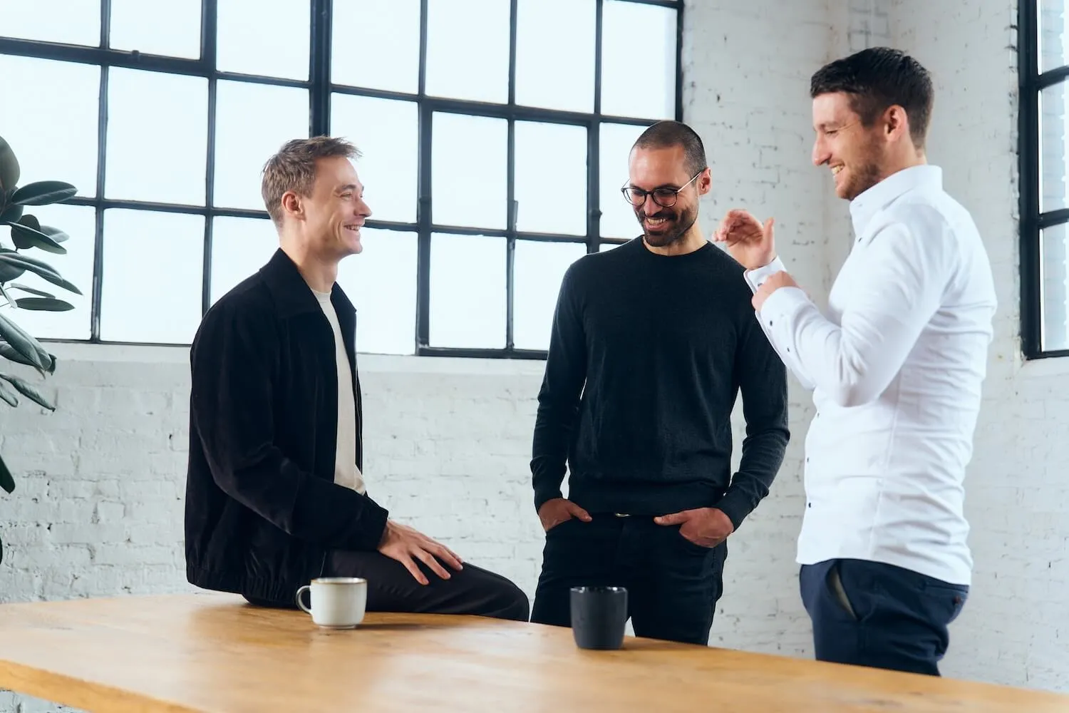 Three men smiling and talking in a bright room with large windows and a wooden table with coffee mugs.