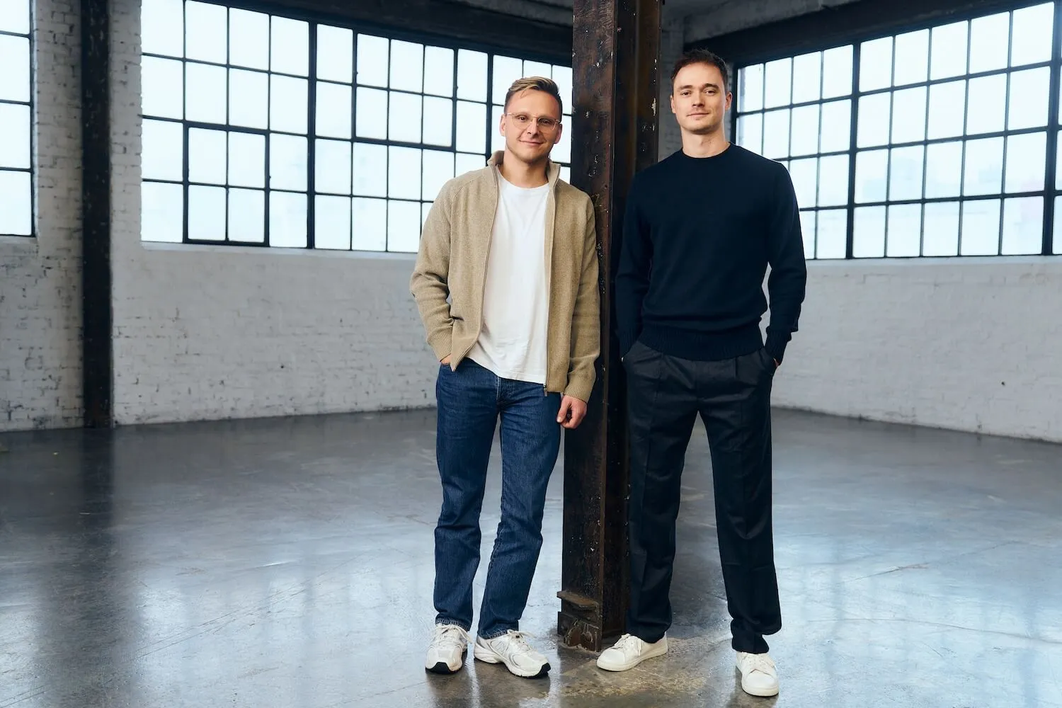 Two men standing casually indoors beside a metal pillar with large grid windows in the background.