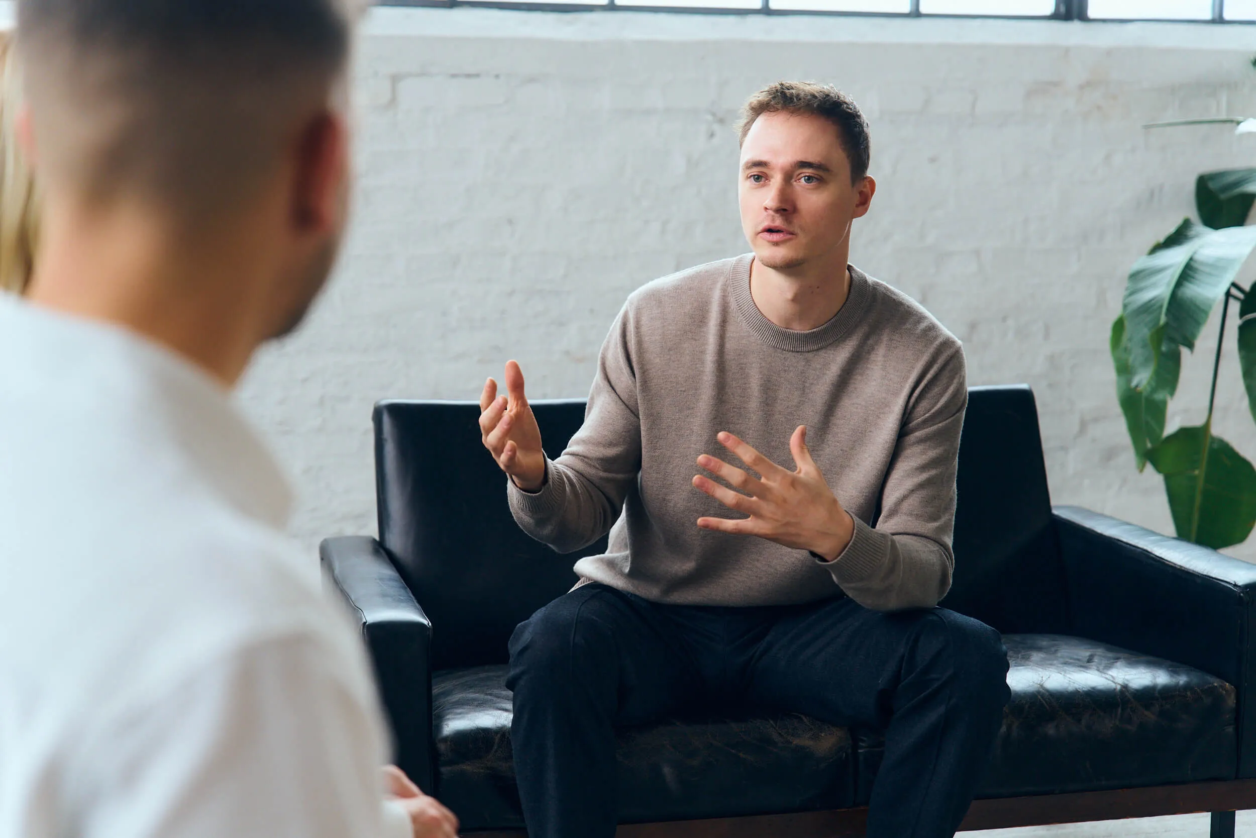 Young man in a brown sweater sitting on a black leather couch gesturing with hands while talking to another person.