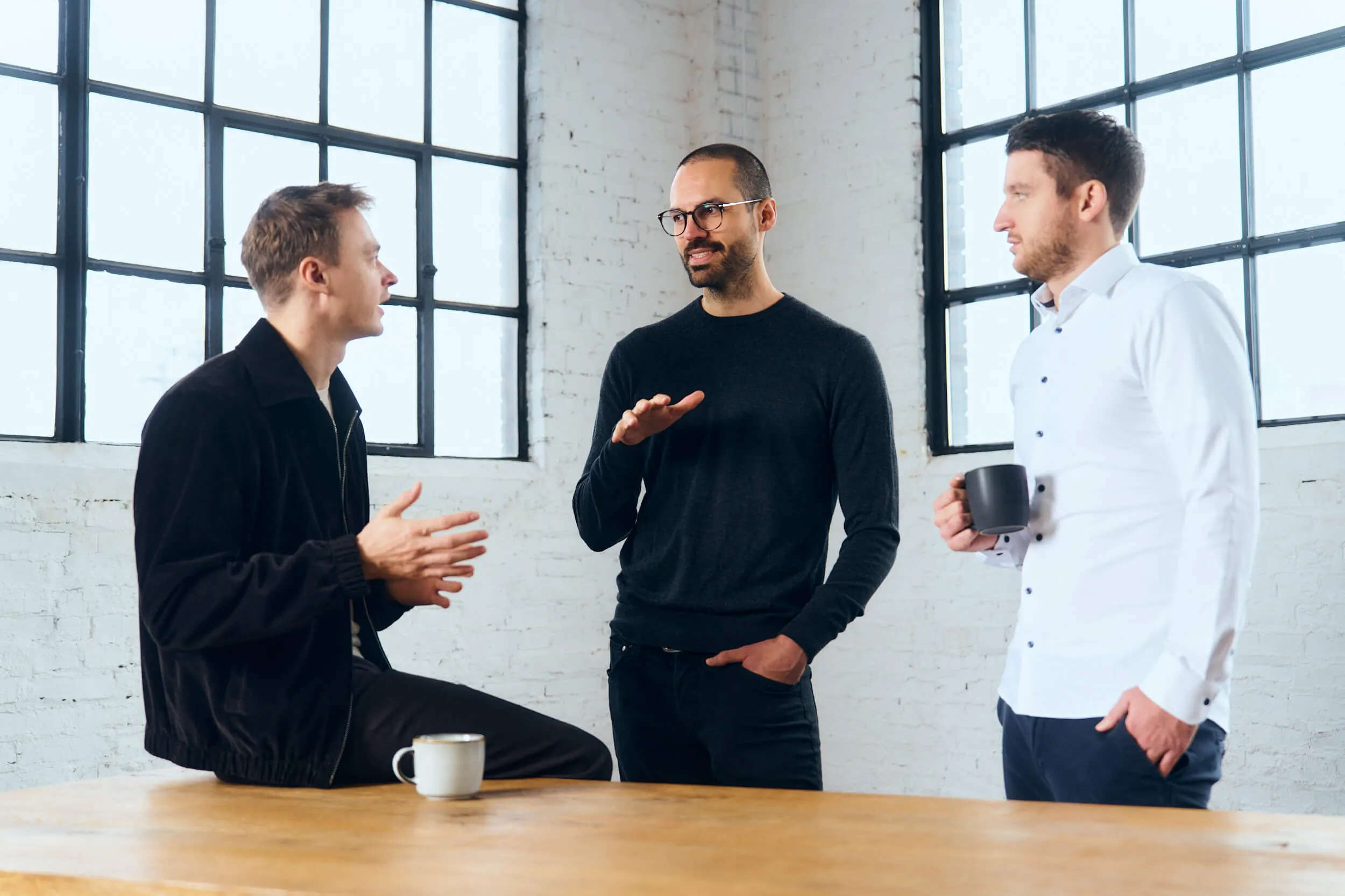 Three men in casual and business casual attire standing by large windows, engaged in conversation with coffee mugs.