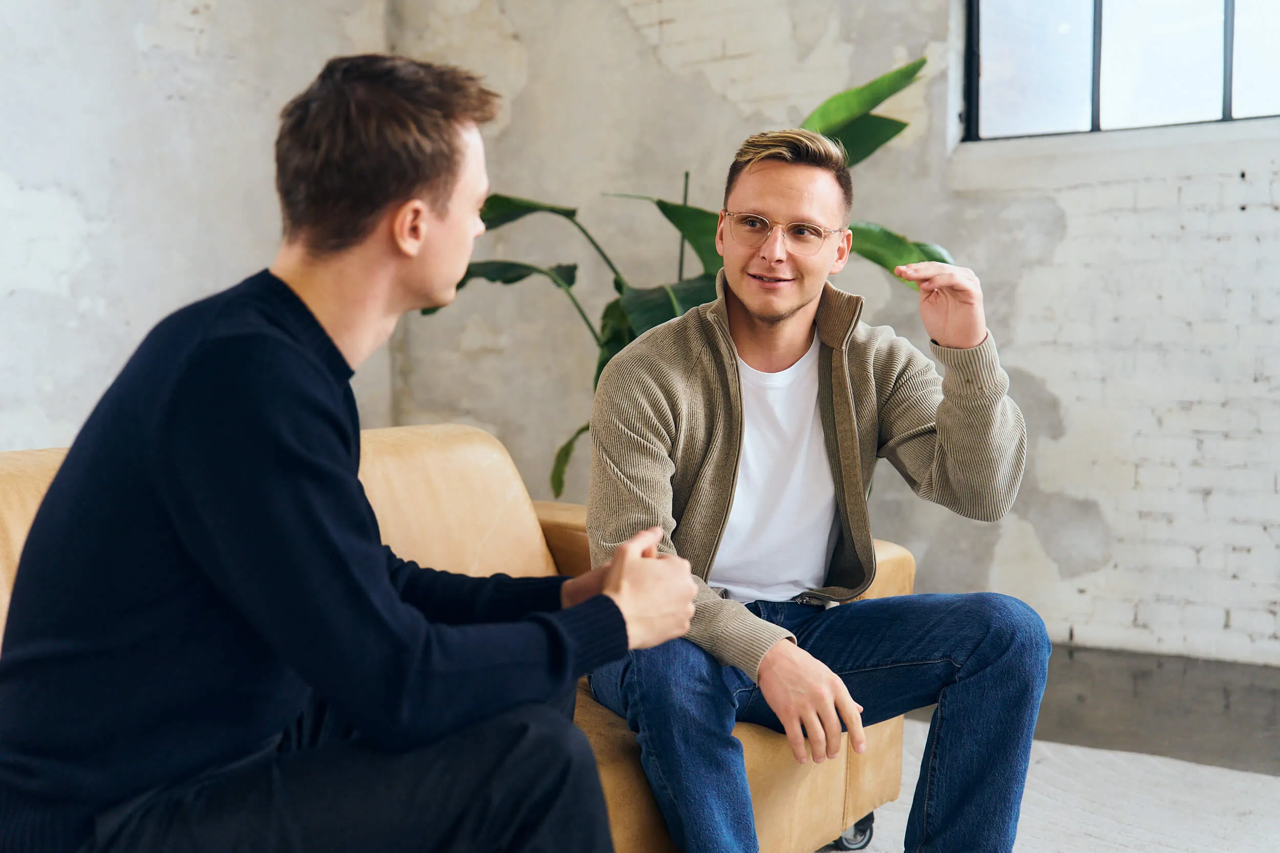 Two men sitting on a tan sofa having a conversation in a bright room with a plant and a window.
