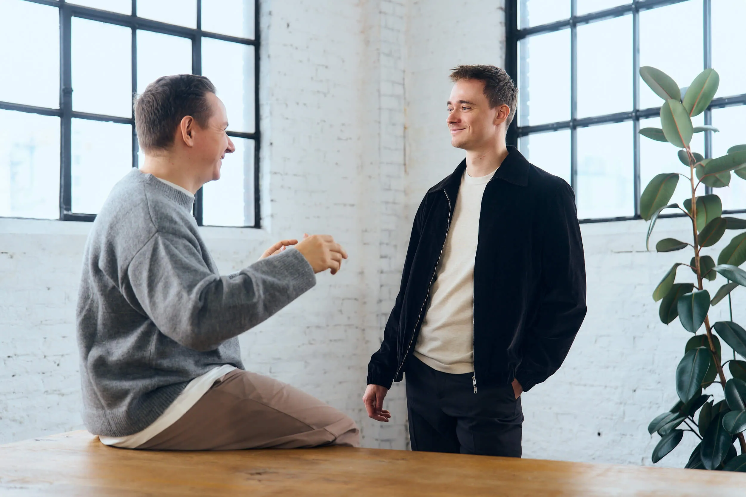 Two men smiling and talking in a bright room with large windows and white brick walls, one sitting on a wooden table and the other standing with hands in pockets.