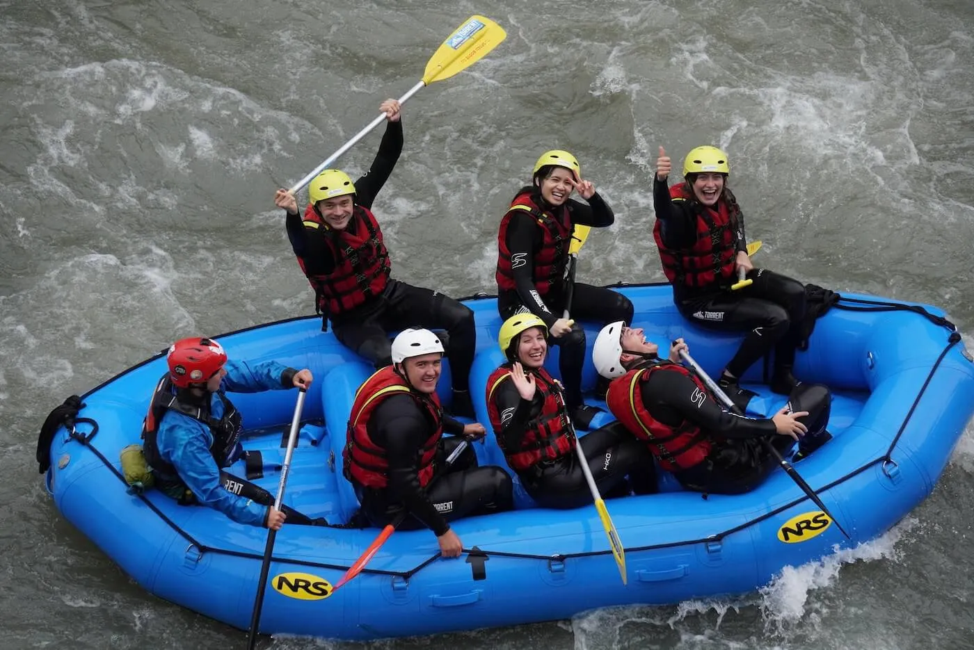 Six people wearing helmets and life jackets happily rafting on a blue inflatable raft in whitewater rapids.