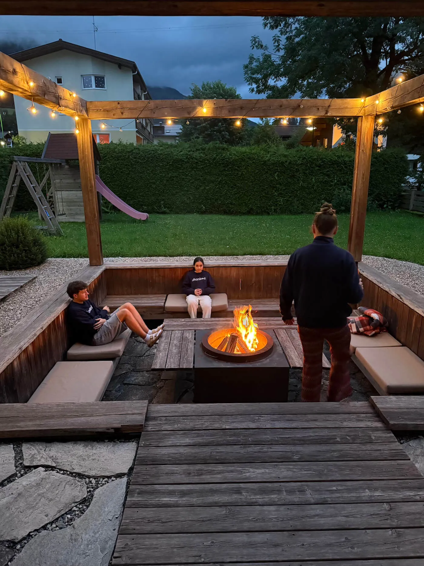 Three people relaxing around a fire pit in a sunken wooden seating area with string lights in a backyard at dusk.