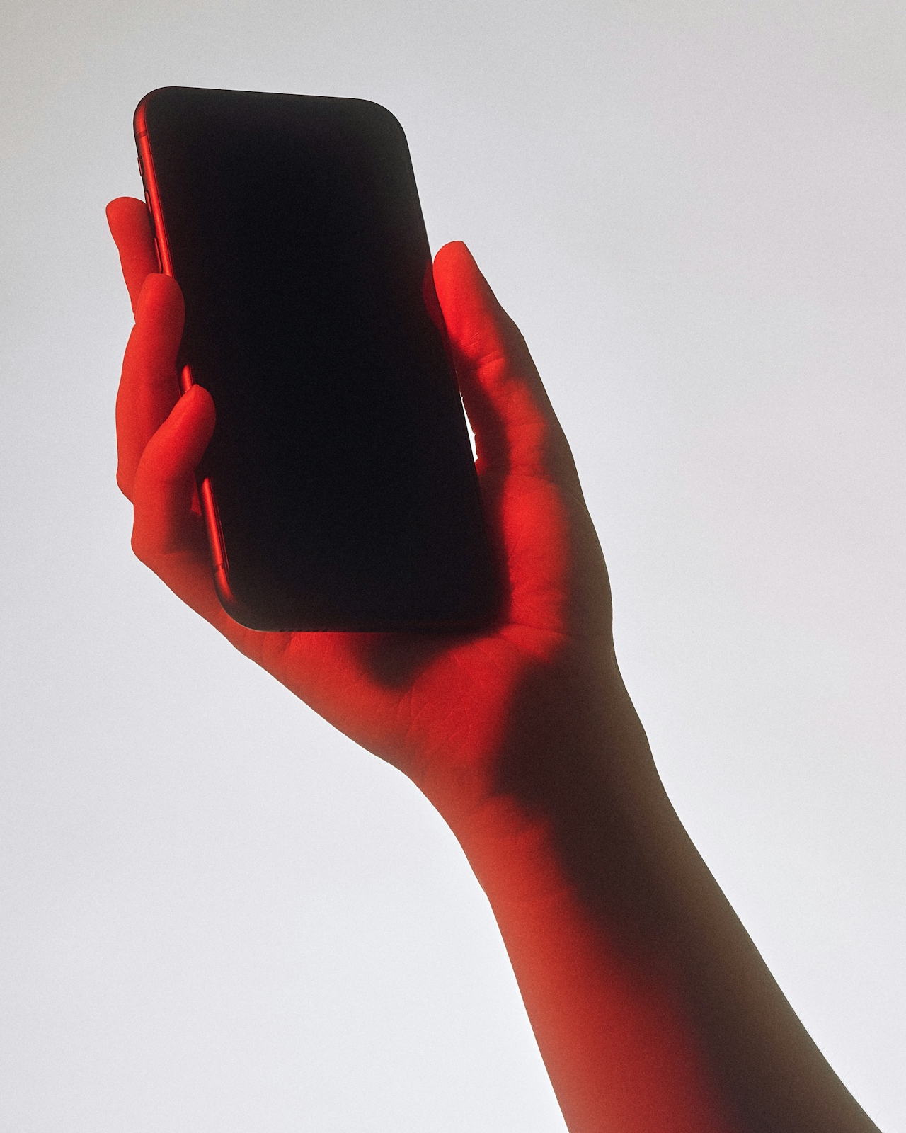 Hand holding a smartphone with red lighting on the skin against a plain background.