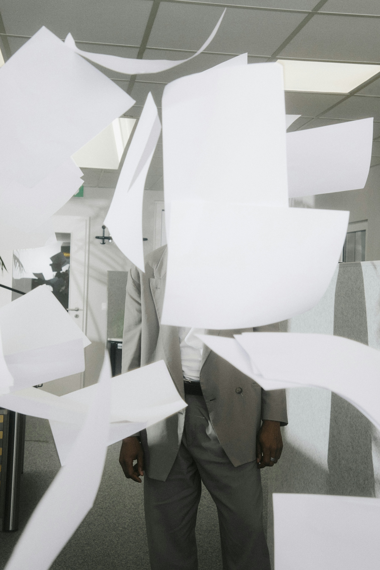 Person in gray suit standing in an office as white papers fly around them, partially obscuring their face.